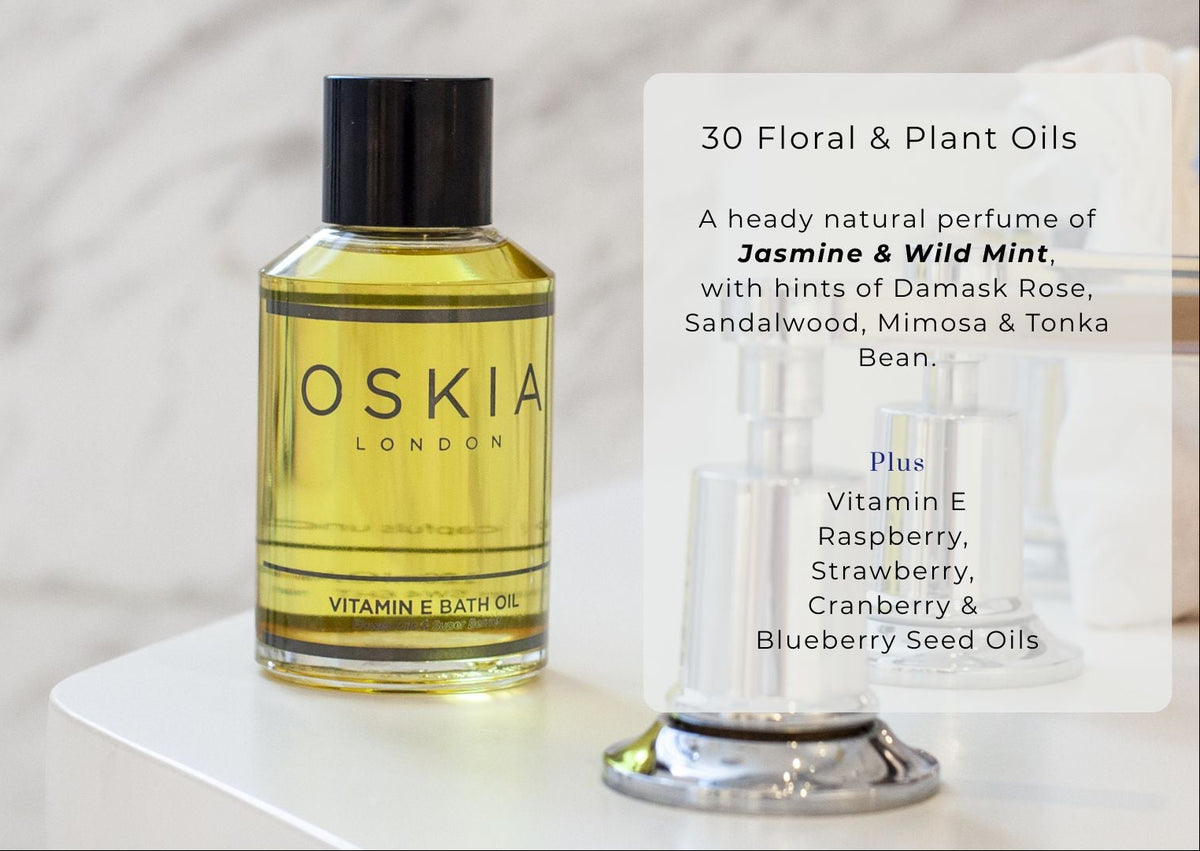 A bottle of Oskia London Vitamin E Bath Oil with golden-yellow oil and a black cap, displayed on a white pedestal next to a clear pump bottle. A text overlay on the right details the scent notes of Jasmine, Wild Mint, and other floral plant oils.